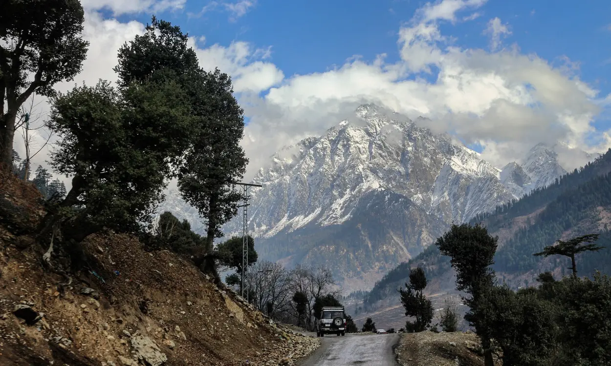 A mountain road through pine trees below snowy peaks in Kalam, Swat Valley, Pakistan